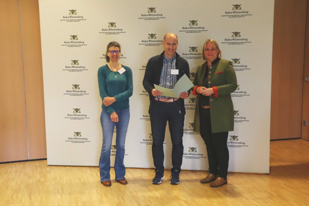 Zwei Frauen und ein Mann stehen nebeneinander. Der Mann hält die Teilnahmeurkunde vor sich.