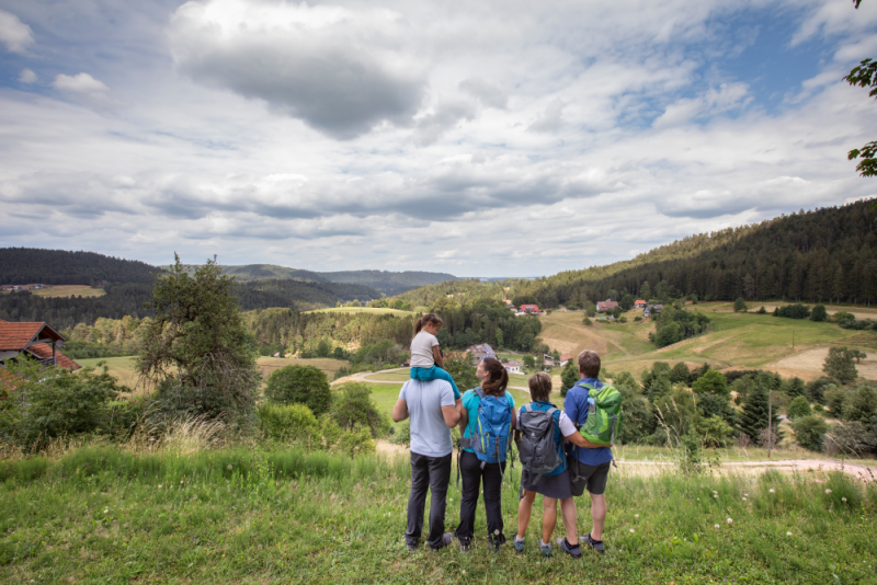 Eine Familie steht auf einer Wiese und schaut auf die Kulturlandschaft mit Wiesen, Weiden, Wäldern und Höfen.