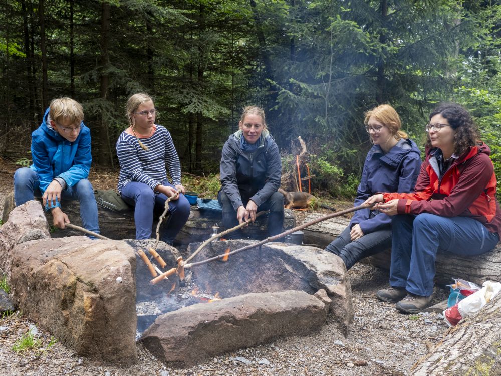 Menschen sitzen zusammen am Lagerfeuer und grillen Würstchen.