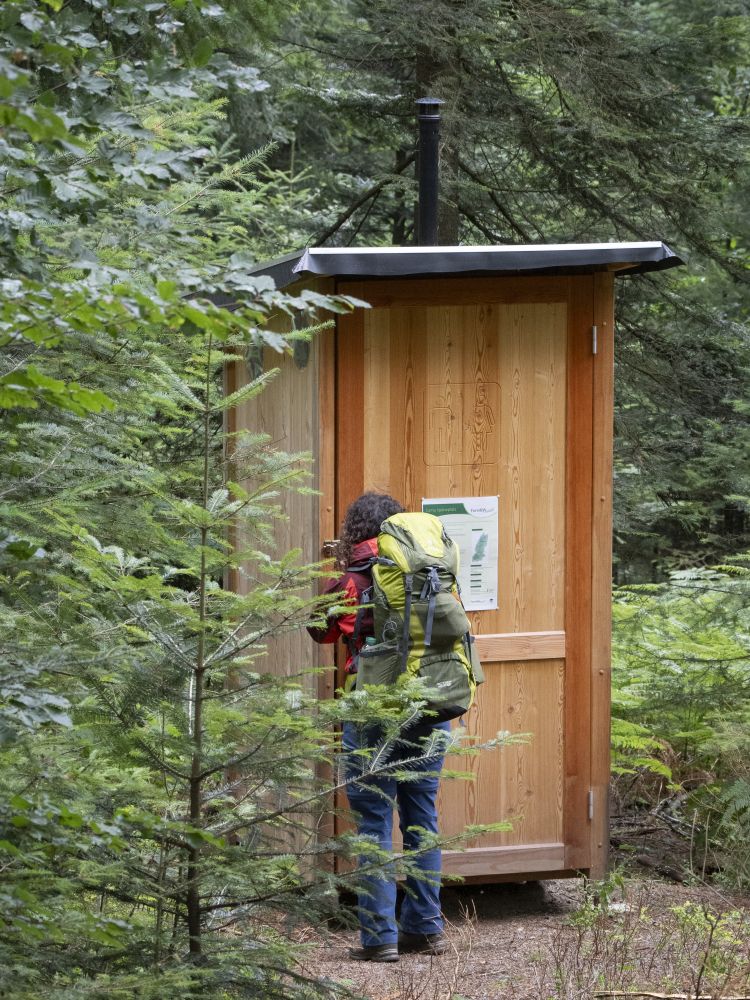 Eine Frau steht vor der Türe eines Holz-Toilettenhäuschens mitten im Wald.