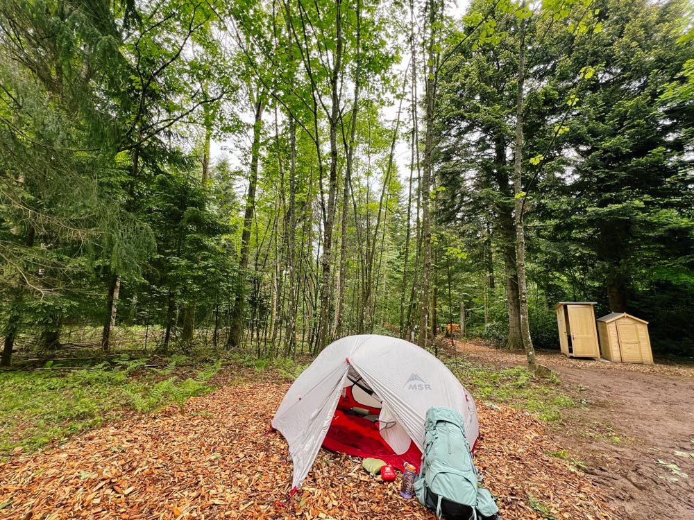 Zelt mit Trekking-Rucksack im Wald. Im Hintergrund ist das Holztoilettenhäuschen zu sehen.