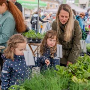 Eine Frau und zwei Mädchen schauen sich Kräuterpflanzen an einem Markt-Stand an.