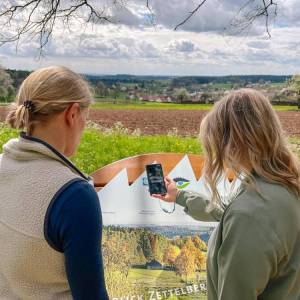 Zwei Frauen stehen vor der Naturpark-AugenBlicke-Panoramatafel in Bad Teinach-Zavelstein und scannen mit dem Handy einen  QR-Code ein.