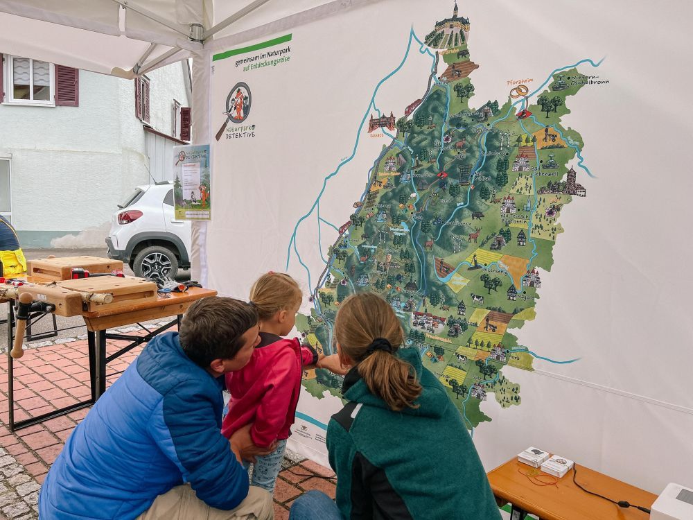 Zwei Erwachsene und ein Kind schauen sich eine große Gebietskarte des Naturparks Schwarzwald Mitte/Nord an.