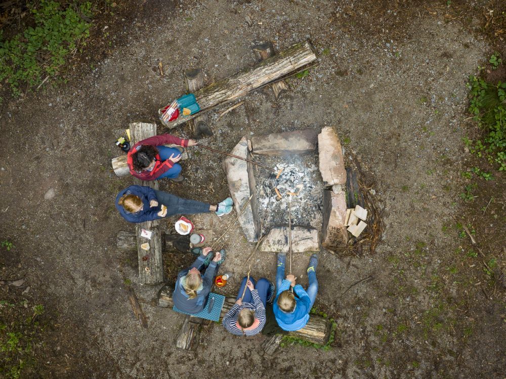 Menschen sitzen an einem Lagerfeuer und grillen Würstchen. Aufgenommen aus der Vogelperspektive.