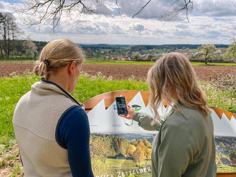 Zwei Frauen stehen vor der Naturpark-AugenBlicke-Panoramatafel in Bad Teinach-Zavelstein und scannen mit dem Handy einen  QR-Code ein.