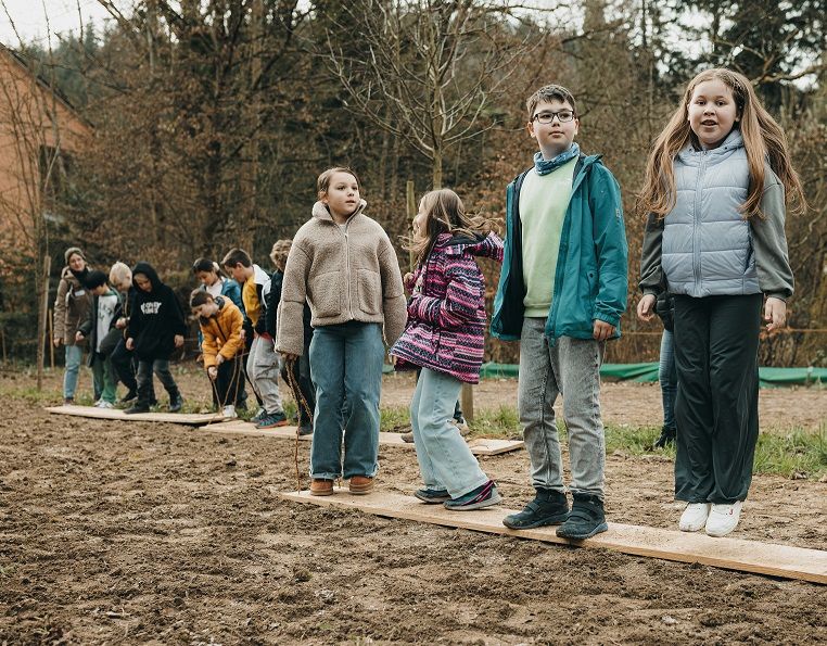 Kinder hüpfen auf Holzlatten auf einer eingesäten Fläche.