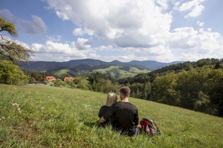 Ein Pärchen sitzt auf einer Wiese und blickt auf die Schwarzwald-Landschaft mit Hügeln, Streuobstwiesen und Wald.