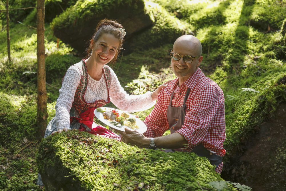 Ein Mann und eine Frau präsentieren ein zubereitetes Gericht in einer Waldkulisse.