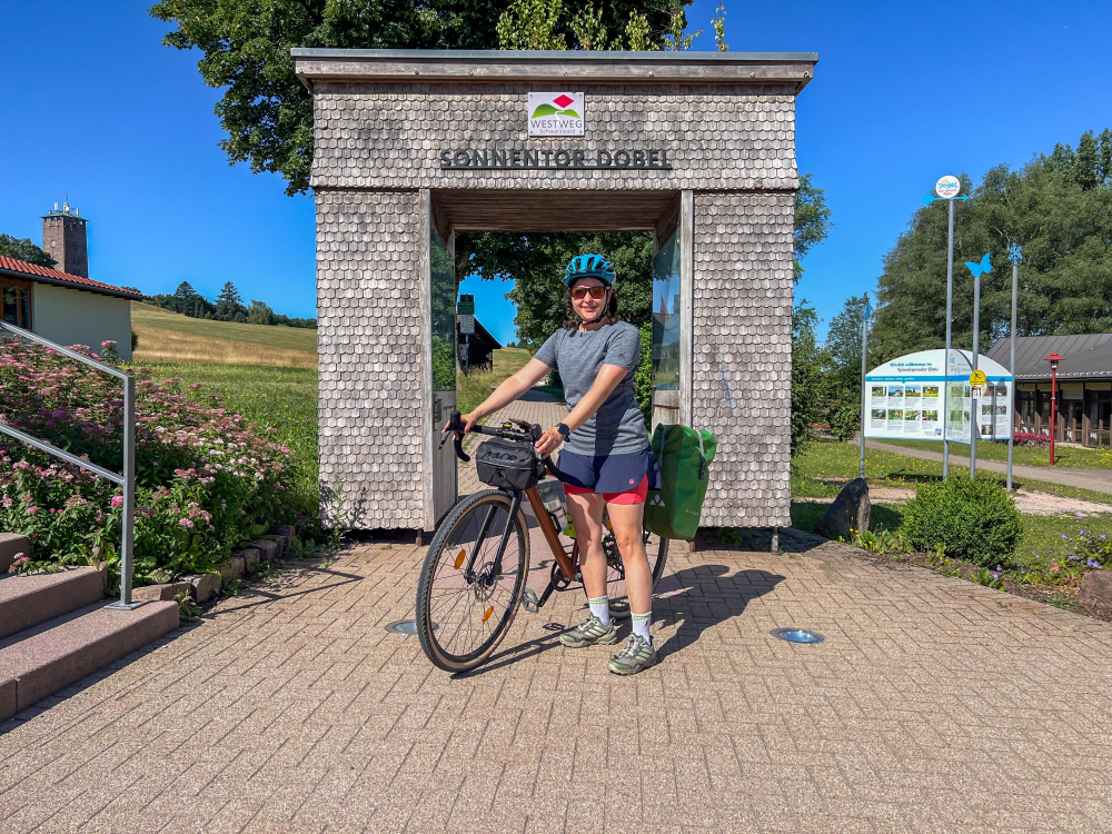 Gravel Bikerin steht vor einem holzernen Torbogen und dem Schild "Westweg".