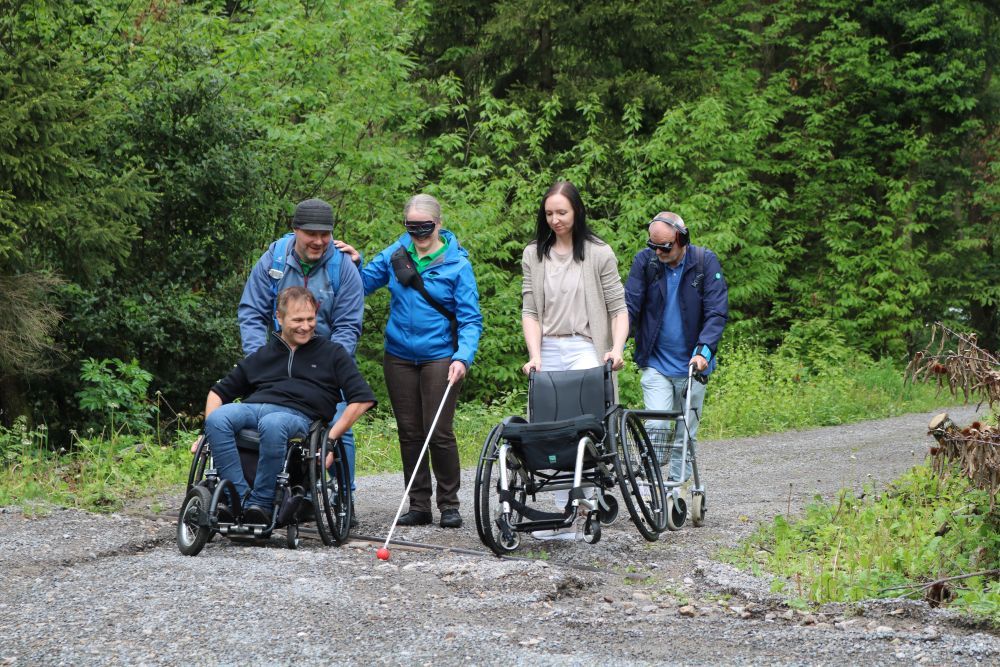 Eine Gruppe von Menschen ist mit Rollstuhl, Blindenstock, Gesichtsmaske und Hörschutz im Wald unterwegs.