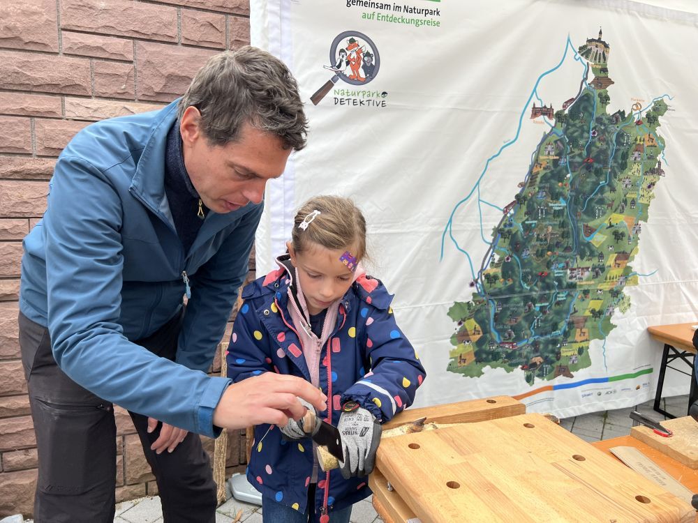 Vater hoilft Tochter an einer Werkbank eine Scheibe von einem Ast abzusägen.