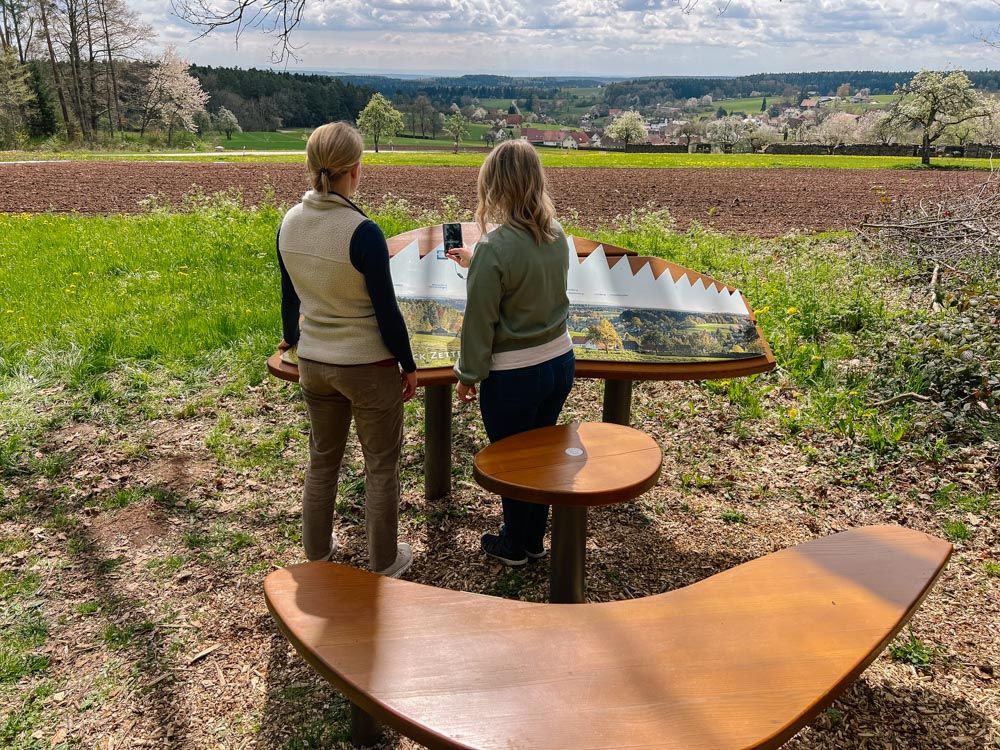 Zwei Frauen stehen beim Naturpark-AugenBlick in Bad Teinach-Zavelstein und scannen einen QR-Code auf der Panoramatafel ein.