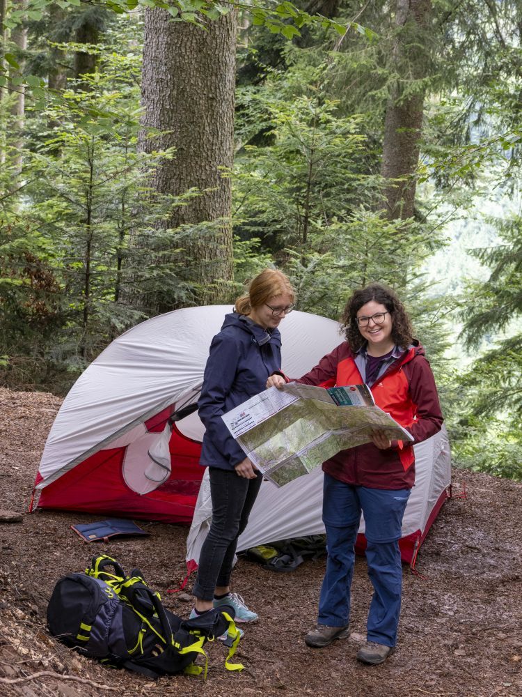 Zwei Frauen stehen vor einem Zelt im Wald und schauen in eine Wanderkarte.