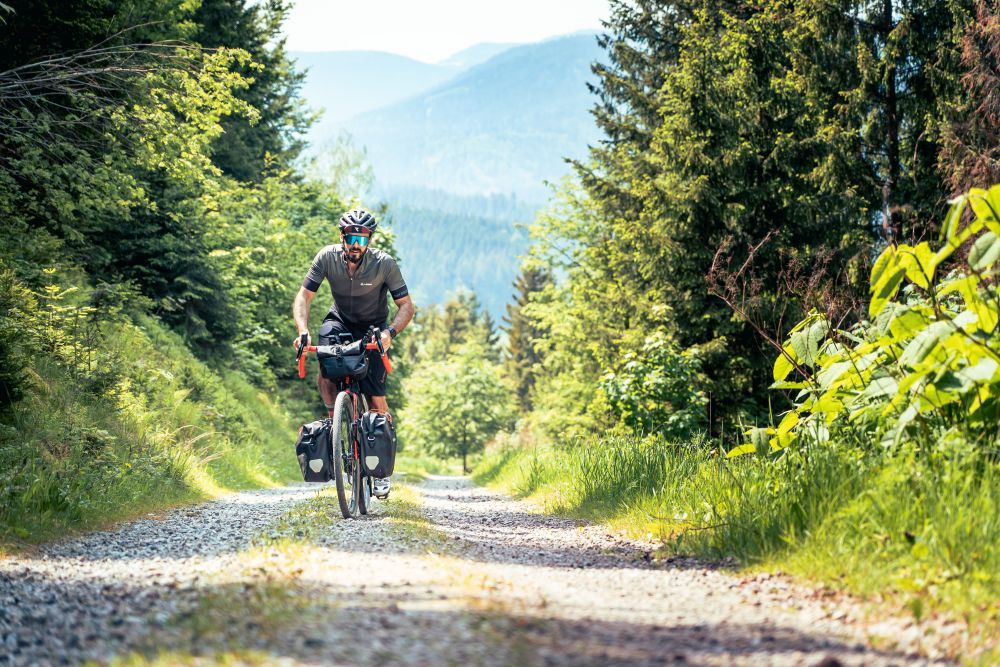 Ein Mann fährt mit einem Gravel Bike einen steilen geschotterten Weg durch einen Wald hinauf.