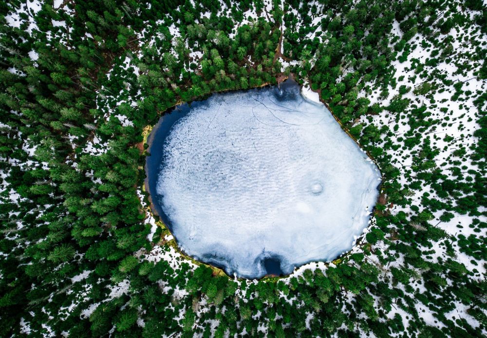 zugefrorener, runder Karsee umgeben von grünen Nadelbäumen