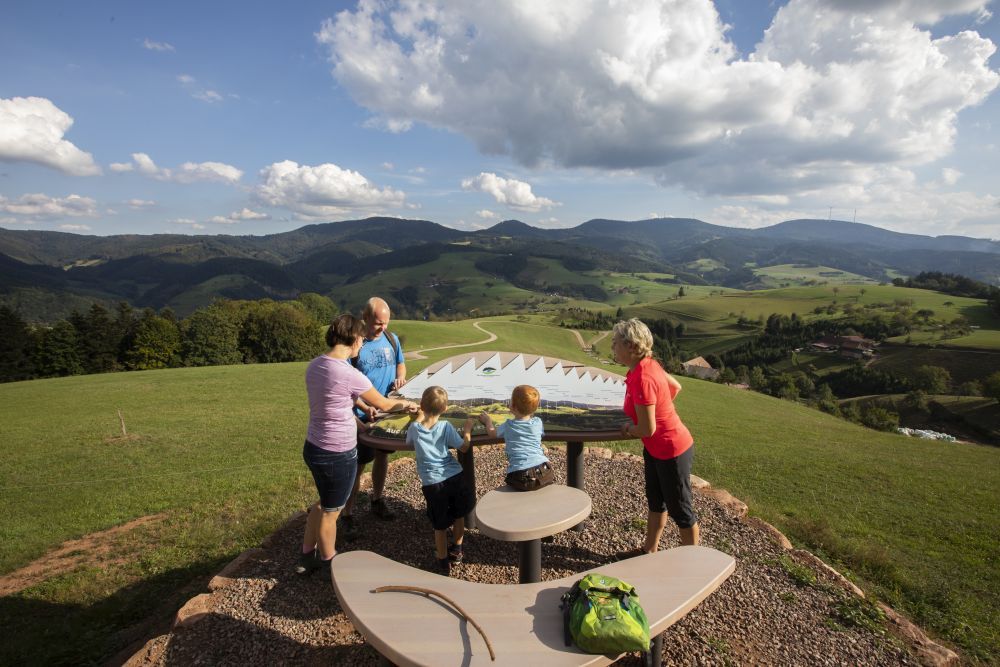 Eine Frau und zwei Kinder schauen auf eine Panorama-Tafel an. Hinter ihnen ist die Kulturlandschaft von Oberharmersbach zu sehen. 