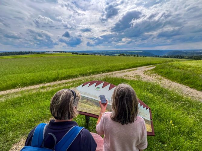 Zwei Frauen stehen vor der Panoramatafel eines Naturpark-AugenBlicks und halten ein Handy vor sich.