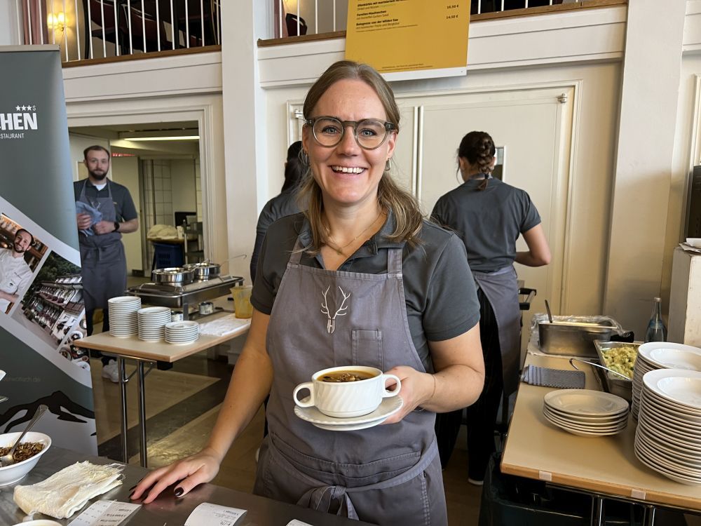 Eine Frau mit Schürze hält eine gefüllte Suppentasse in der Hand.