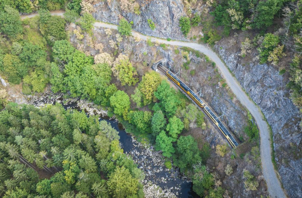 Ein Fluss verläuft im Wald, am felsigen Bergrand fährt eine Bahn.